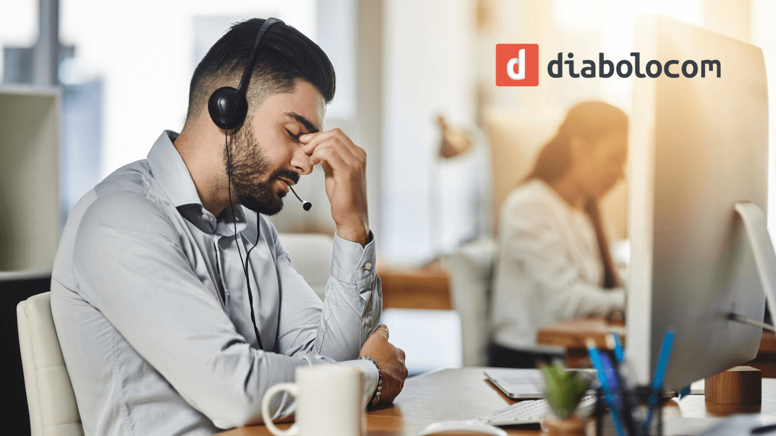 Stressed contact centre agent wearing a headset pinches the bridge of his nose at his desk, with a colleague working in the background and the Diabolocom logo top right.