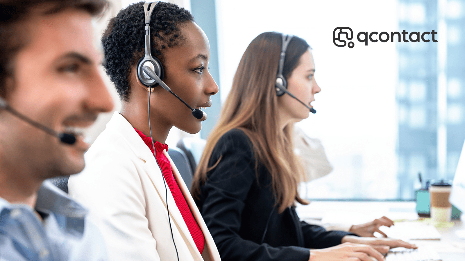 Three contact centre agents wearing headsets work at computers in a bright, modern office with city views behind them. The qcontact logo appears in the top right corner.