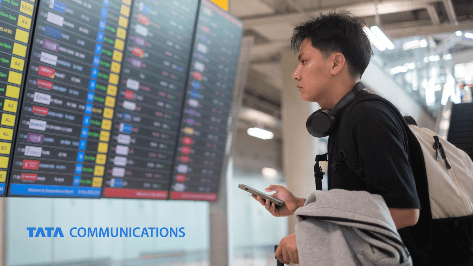 Young traveller with headphones and a backpack holding a smartphone while looking up at an airport departures board, with the Tata Communications logo in the bottom left corner.