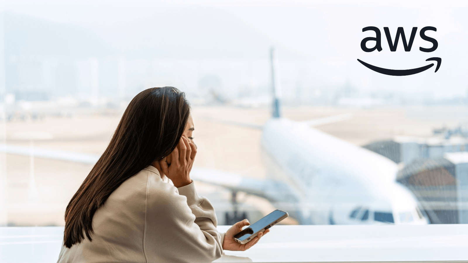A woman rests her head on her hand while holding a smartphone, gazing out of an airport window at a parked plane, with the AWS logo top right.