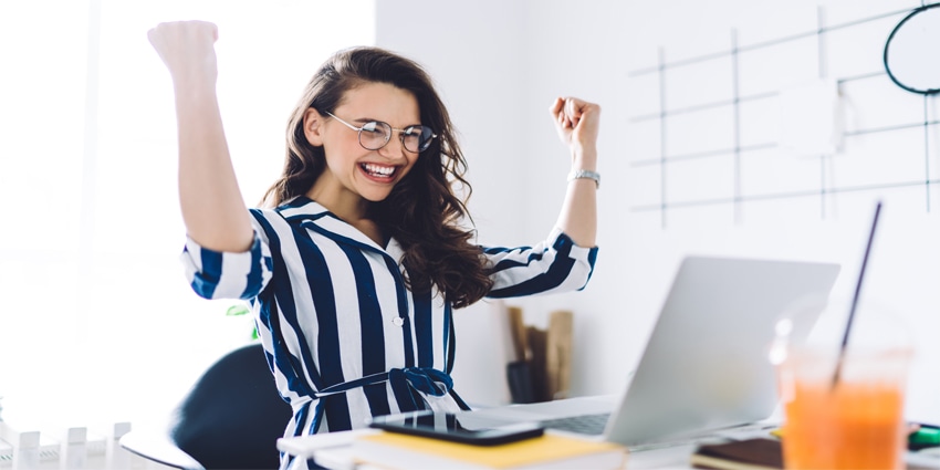 Excited young woman sitting at table with laptop and celebrating success