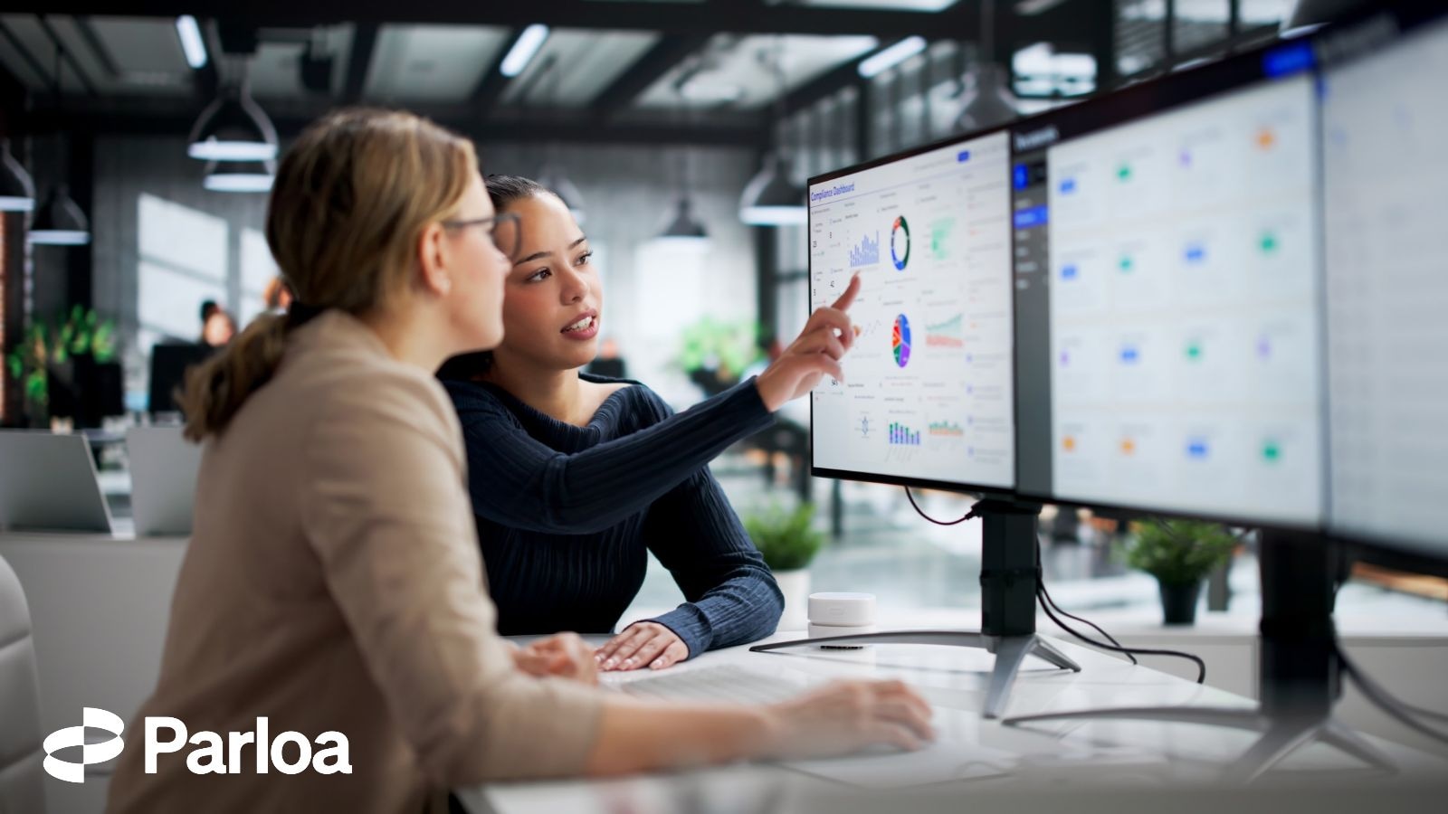 Two colleagues in an office review a dashboard with charts and performance data on a large monitor, one pointing at the screen as they discuss it together
