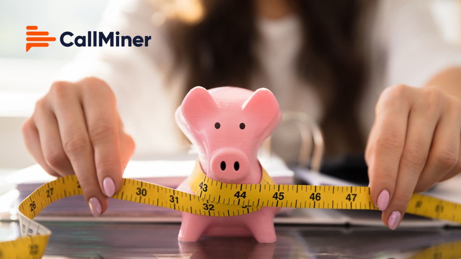 A person wraps a yellow measuring tape around a pink piggy bank on a desk, with the CallMiner logo in the top left corner